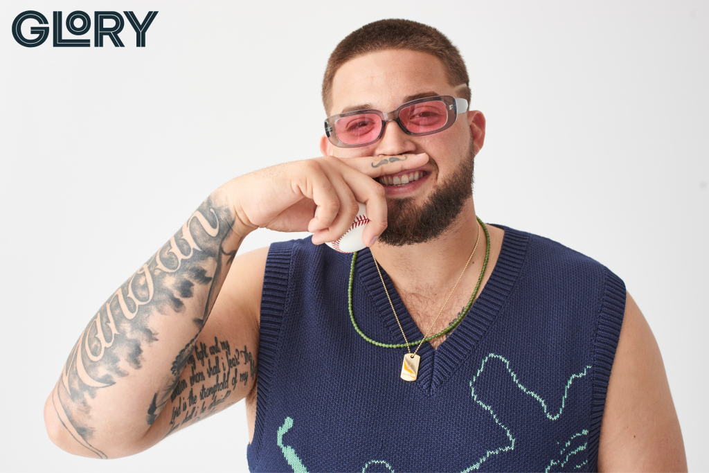 Alek Manoah in pink sunglasses holding finger to lip with mustache tattoo wearing blue sweater vest