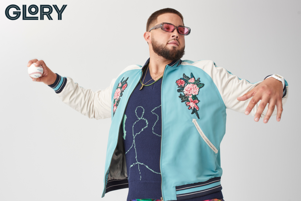 Alek Manoah wearing pink tinted sunglasses, dark blue knitted vest under a silk bomber jacket with florals and mock pitching a baseball